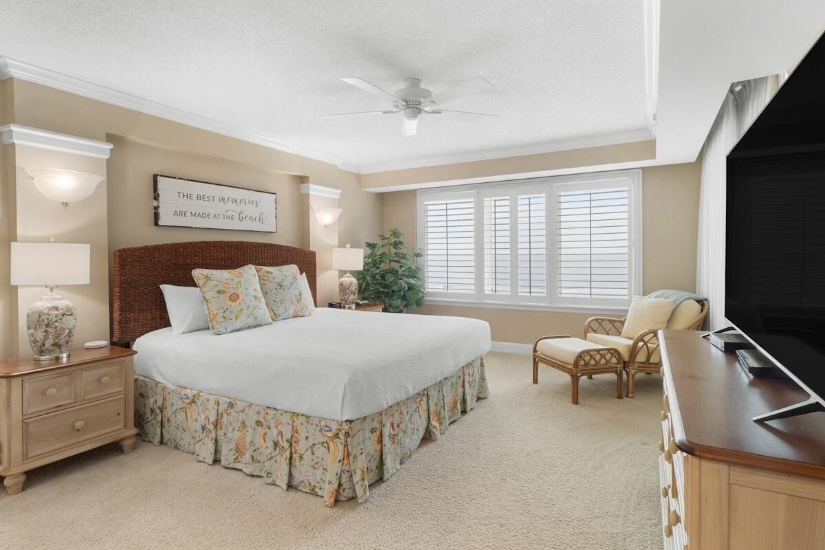 A bedroom with a large bed, a ceiling fan, a window with blinds, a nightstand with a lamp, and a television.