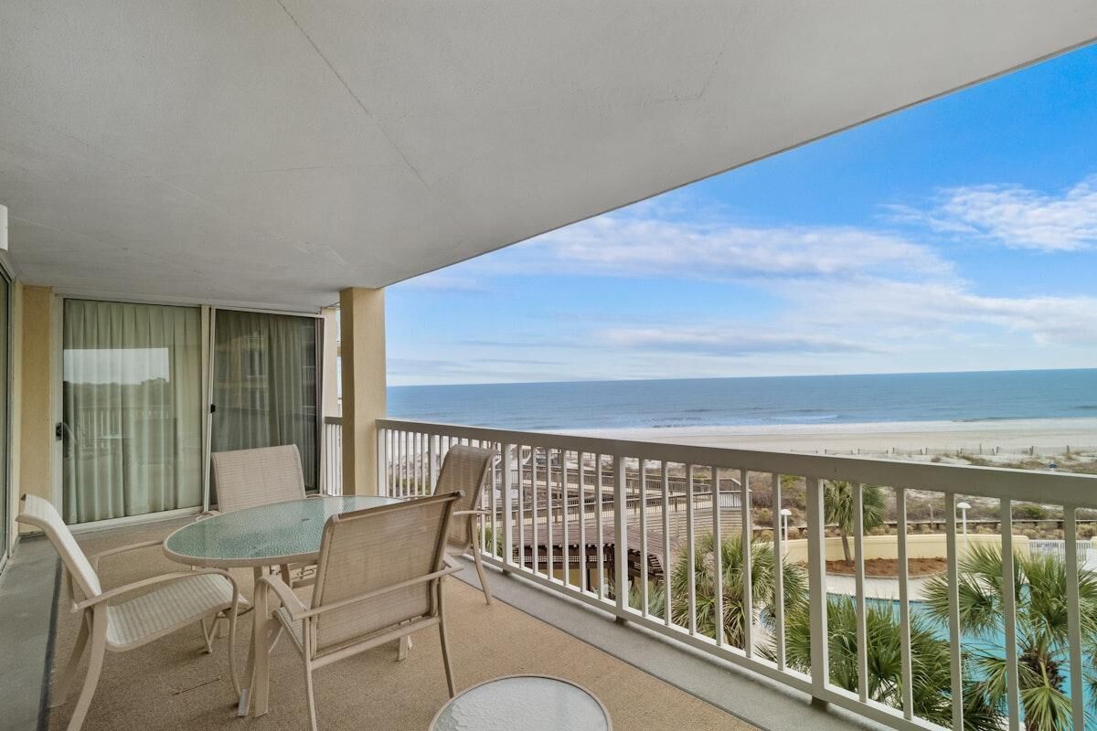 A balcony with a table and chairs, overlooking a beach and a pool.