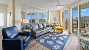 Un vaste salon comprenant un canapé en cuir gris, un fauteuil bleu et un fauteuil jaune. Une grande fenêtre offre une vue sur les bâtiments, et un balcon est aménagé avec du mobilier d’extérieur.