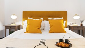 A neatly made bed with a headboard, two bedside tables with lamps, and a plate of croissants and two cups on the bed.