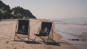 Two beach chairs with "2-HOME" branding on a sandy beach with a calm sea and a forested hill in the background. - Laguna Basen SPA Przy pla y Gara Wifi (Mielno)
