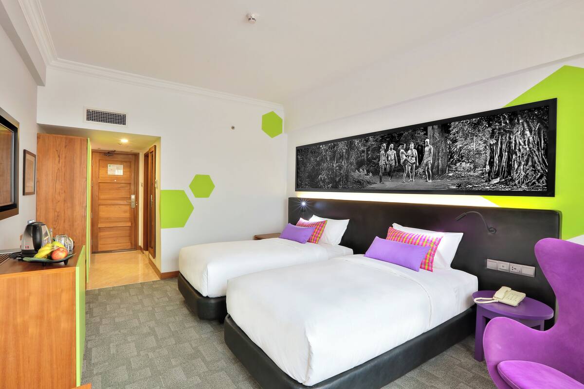 A hotel room with two beds, a desk with a kettle, a chair, and a large painting on the wall.