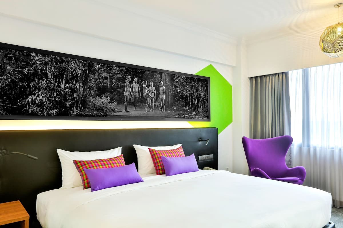 A hotel room with a large bed, a headboard, a wall-mounted artwork, a purple chair, and a window with curtains.