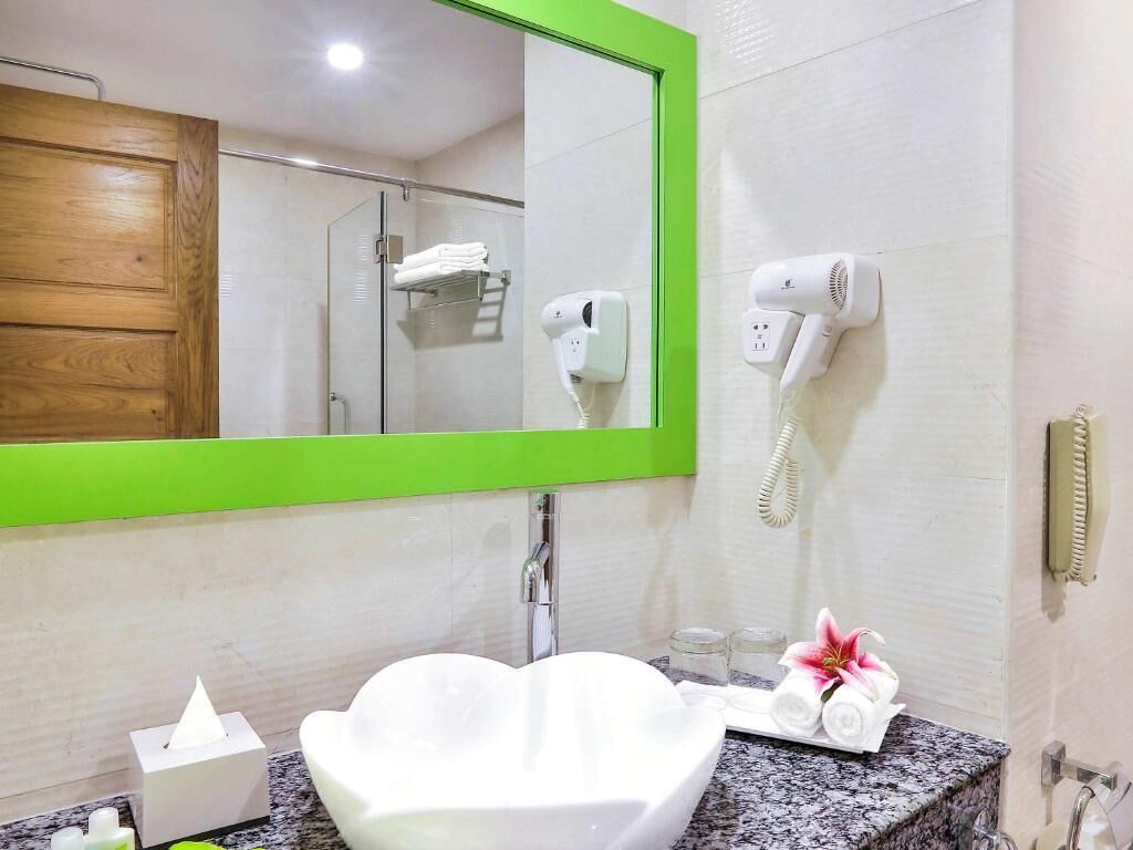 A bathroom with a heart-shaped sink, a green-framed mirror, a hairdryer, and a towel rack.