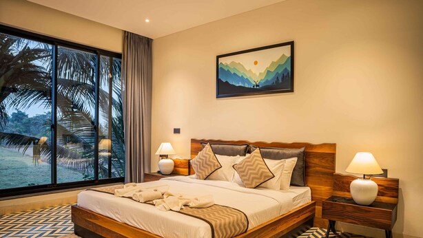 A spacious bedroom with a large bed, wooden headboard, and a view of palm trees through floor-to-ceiling windows. - Marvelous villa with AC in Saligao - 5 bedrooms available (Saligao)