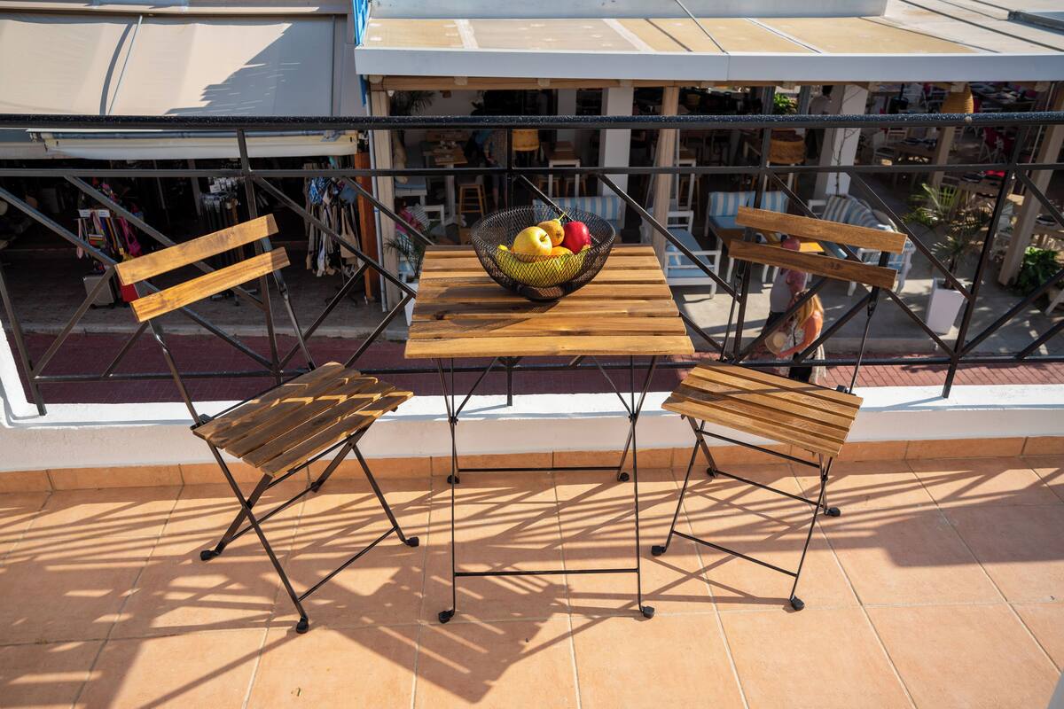 Um terraço com mesa de madeira e duas cadeiras, guarda-corpo de metal e uma cesta de frutas.