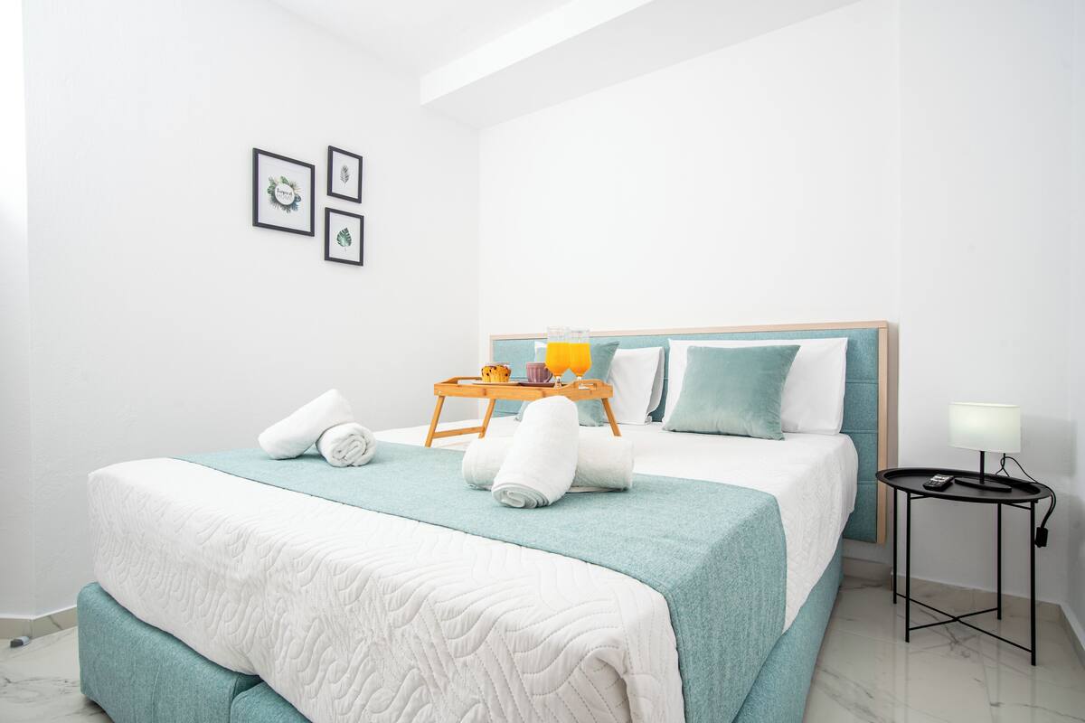 Cama bem arrumada com cobertura verde-água e branca, mesa de cabeceira com abajur e bandeja com toalhas e caneca.
