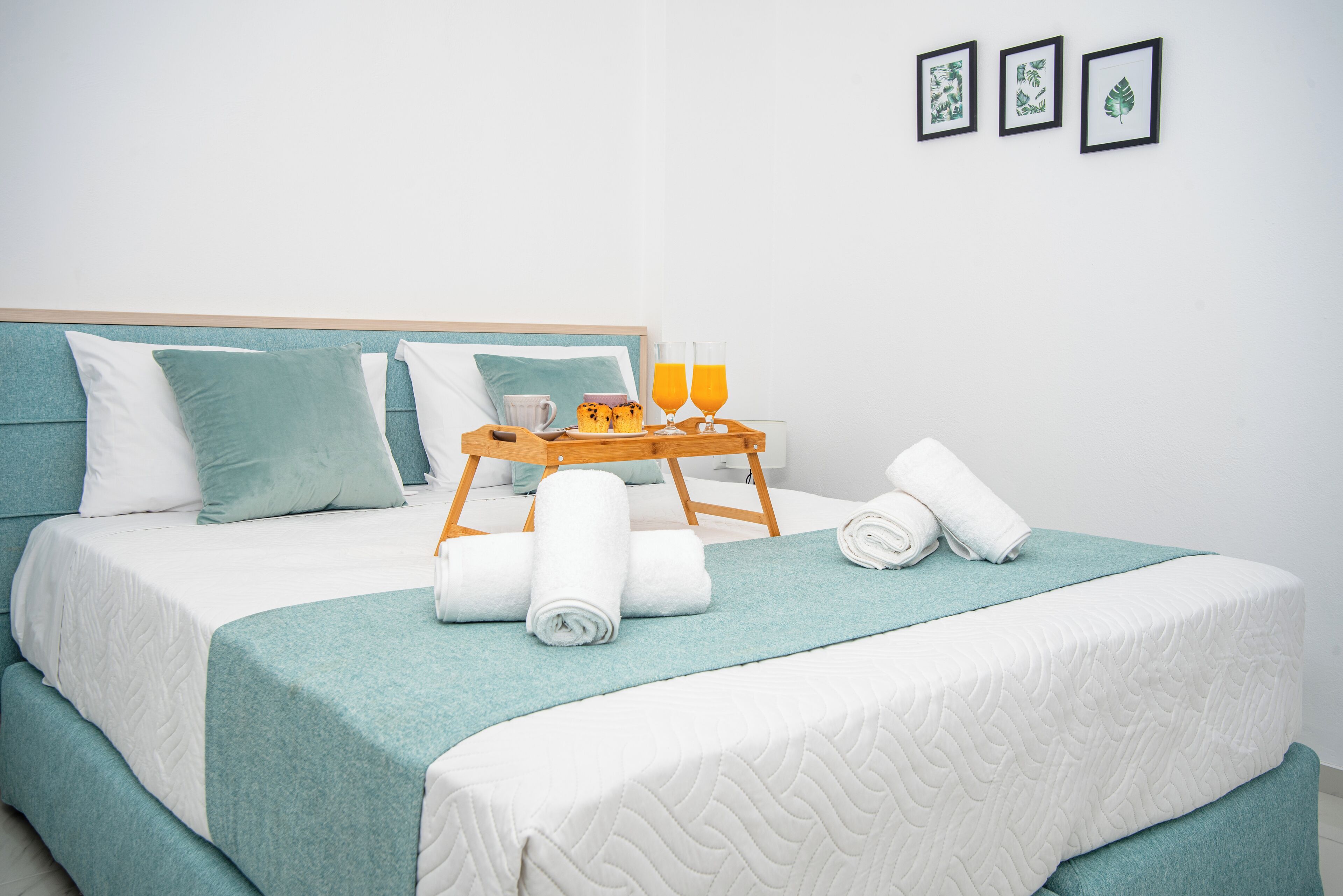 Un lit bien fait, recouvert d’une courtepointe turquoise, avec des oreillers blancs et un plateau en bois sur lequel se trouvent une tasse, des pâtisseries et deux verres de jus d’orange.