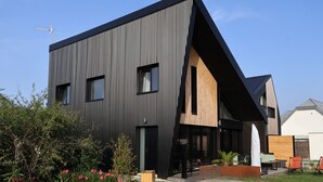 A modern two-story house with a sloped roof, a wooden deck, and a gravel driveway.