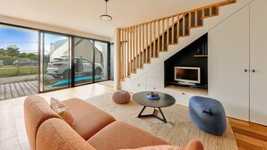 A modern living room with a sofa, ottoman, and a round coffee table. A staircase with wooden railings leads to a lower level.