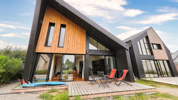 A modern house with a wooden facade, large glass windows, and a deck with outdoor seating.