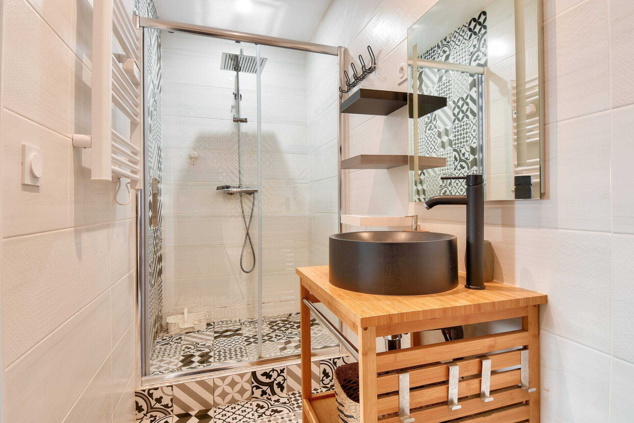 Une salle de bain moderne avec une douche à parois de verre, un meuble-lavabo en bois avec un lavabo noir, des carreaux à motifs et un miroir décoratif.