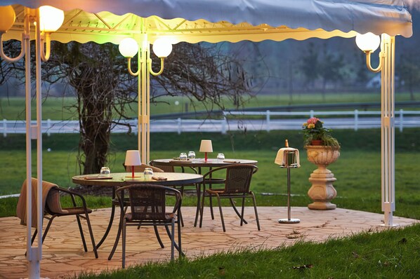 An outdoor patio area with round tables and chairs, a covered seating area with lights, and a decorative urn. - AUBERGE DU PORT (Remilly-Aillicourt)