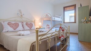 A well-lit bedroom with a bed, two heart-shaped pillows, a metal-framed bed, a nightstand, a lamp, a bunk bed, a window with blinds, and a green cabinet with glasses and a bottle. - Wavefront San Foca (Melendugno)