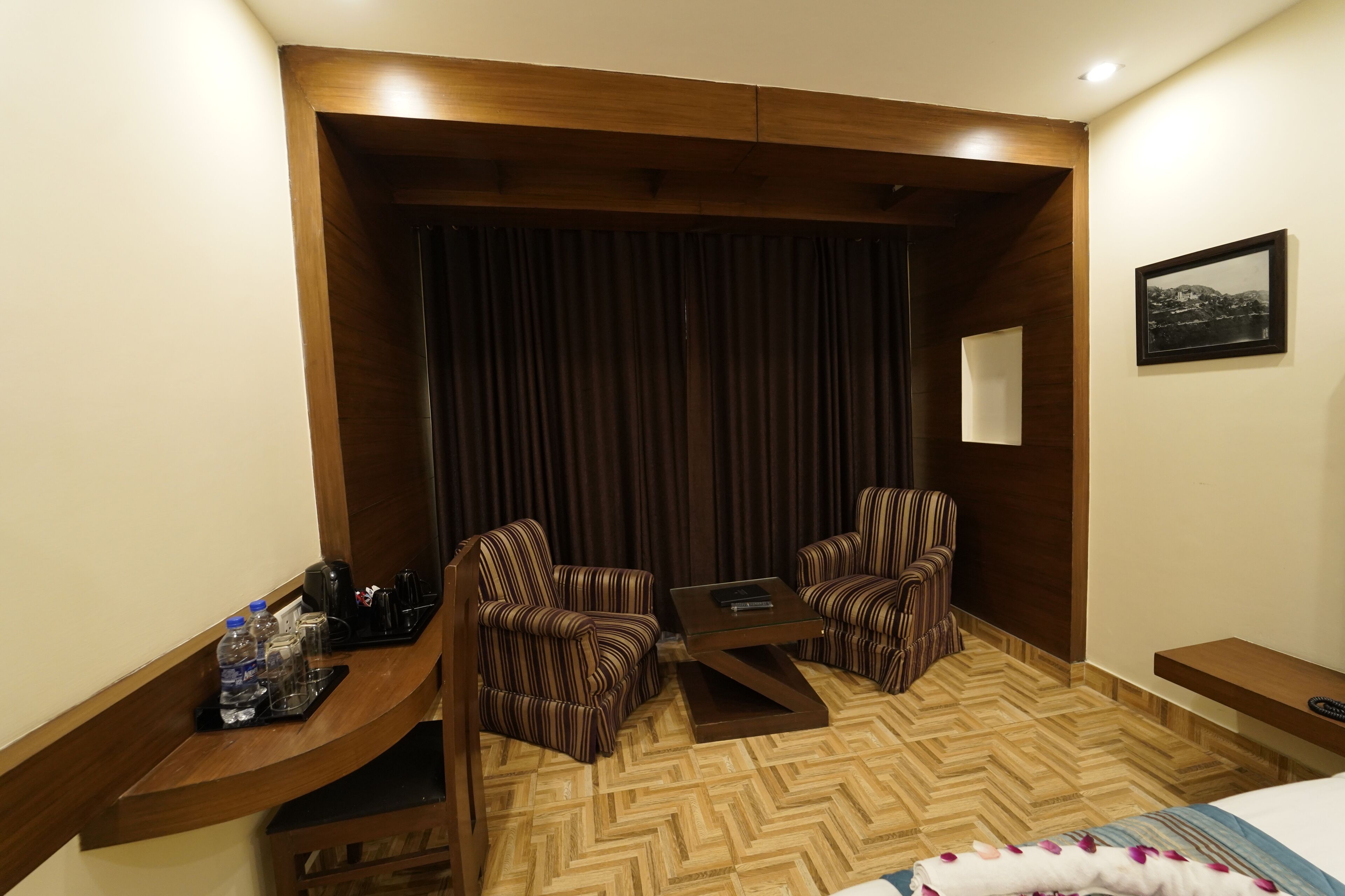 A hotel room with a wooden accent wall, a large window with curtains, two striped armchairs, a small table, and a bed with a striped blanket.