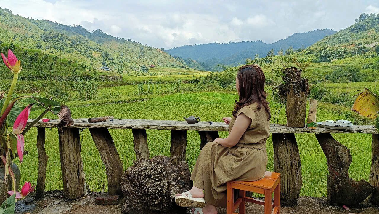 Una mujer sentada en un banco de madera contemplando un paisaje verde exuberante con campos en terrazas y montañas al fondo.