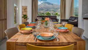 A dining area with a wooden table set for six, surrounded by yellow chairs, with a mountain view through large windows. - Villa Hemèra, pool and relaxation a few minutes from Scopello (Sicilia)