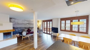 A modern living room with a fireplace, a television, and a dining area with wooden chairs and a table. - Reka-Ferienanlage Rougemont (Gstaad) (Rougemont)