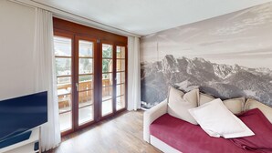 A room with a wooden floor, a TV on a stand, a sliding glass door leading to an outdoor balcony, and a large mountain landscape mural on the wall. - Reka-Ferienanlage Rougemont (Gstaad) (Rougemont)