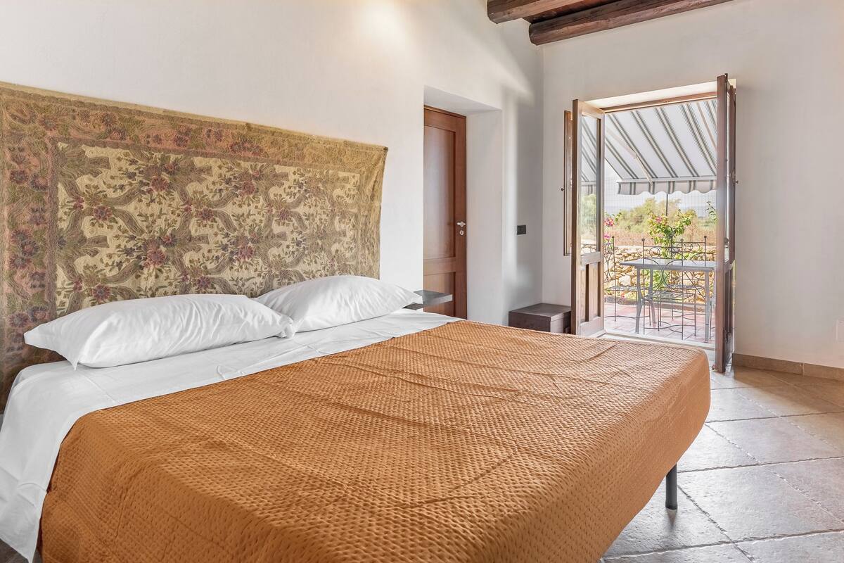 Un dormitorio con una cama grande, un cabecero con un tapiz floral y vistas a un balcón con mobiliario exterior.