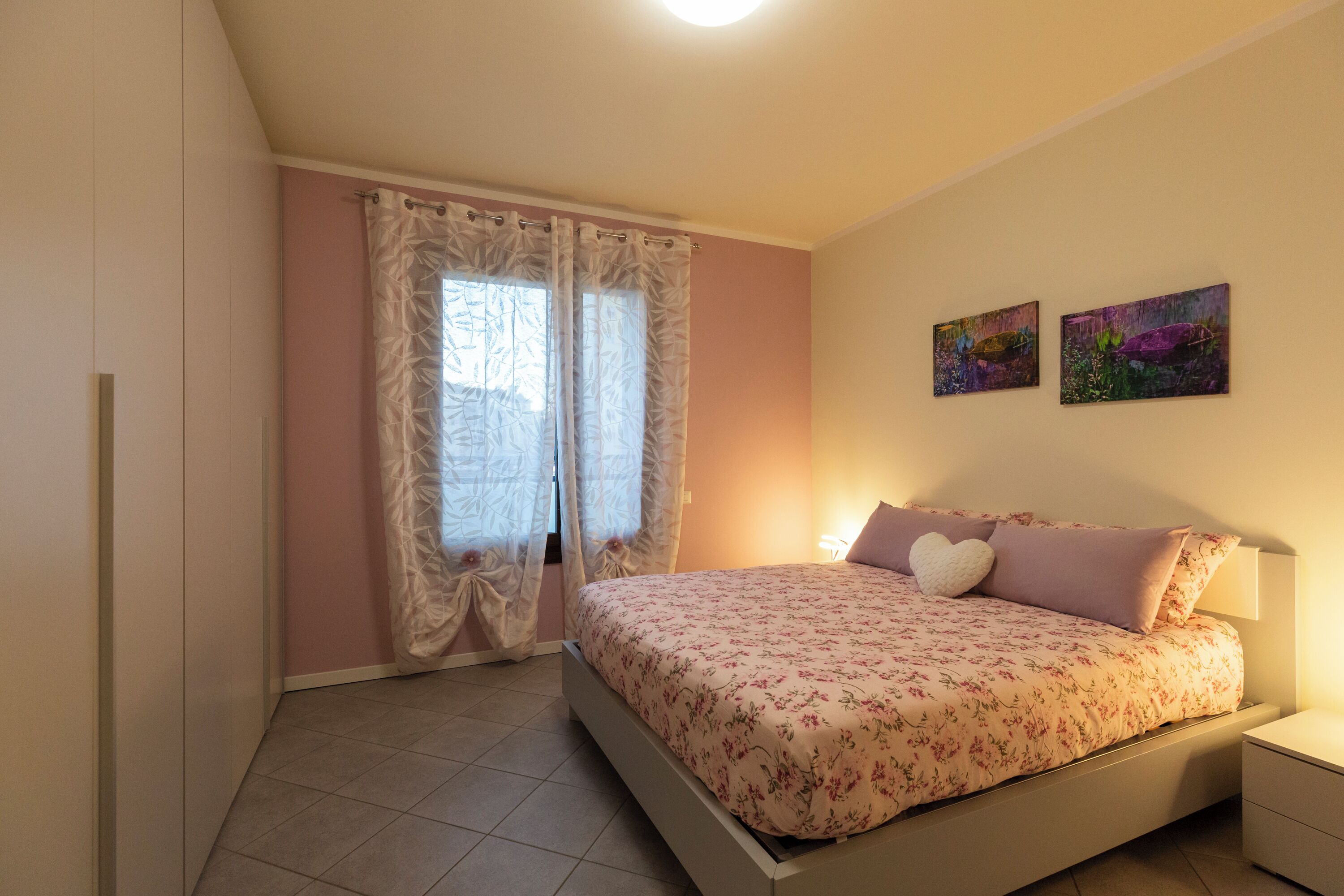 Une chambre à coucher avec un lit, des rideaux, deux tableaux accrochés au mur et une table de chevet.