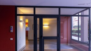 A modern building entrance with glass doors, a red wall, and a visible hallway. - notaMi - UniGarden - Bicocca District (Milan)