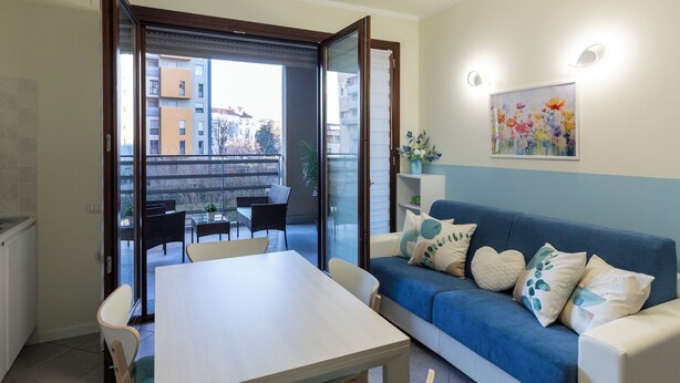A modern living room with a blue sofa, white table, and chairs. A sliding glass door leads to an outdoor balcony with a dining area. - notaMi - UniGarden - Bicocca District (Milan)