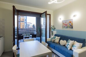 Un moderno salón con un sofá azul, una mesa blanca y sillas. Unas puertas corredizas de vidrio dan a un balcón exterior con zona de comedor.