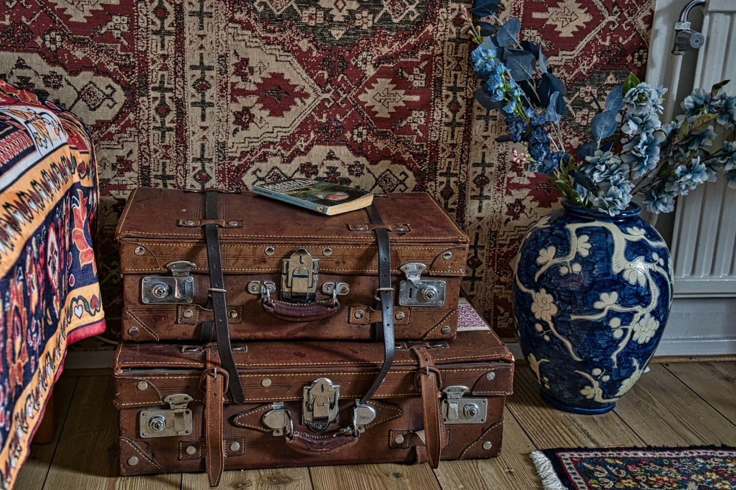 A stack of brown leather suitcases with metal clasps, a book, and a blue and white vase with flowers on a wooden floor.