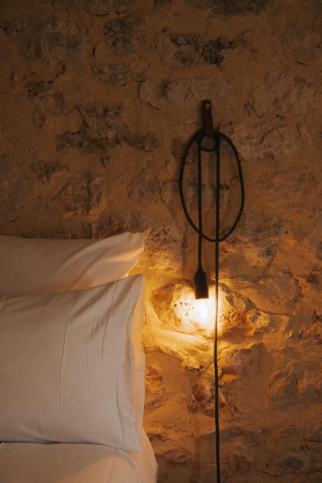 Un lit dans une chambre dont un mur est recouvert de pierres apparentes, et où l’on trouve un simple luminaire mural au style rustique.