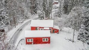 A red house with a snow-covered roof and a white garage door, surrounded by snow-covered trees and a road. - Nice home in Hovsherad with WiFi (Hovsherad)