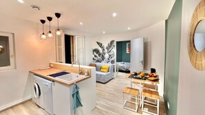A compact living space with a kitchenette, a washing machine, a dining area with a table and chairs, and a bed in the background. - Le Victor Hugo - Clichy-la-Garenne (Clichy)