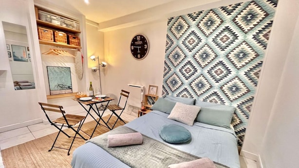 A modern bedroom with a bed, a small table with chairs, a wall clock, and a patterned headboard.