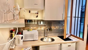 A modern kitchen with a stainless steel sink, a kettle, and a microwave. There are wine glasses on a shelf, and a window with a view of the outside.