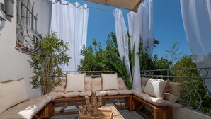 Un espace extérieur lumineux, aménagé avec des sièges, comprenant un banc rembourré et une petite table, le tout entouré de rideaux blancs et de verdure.