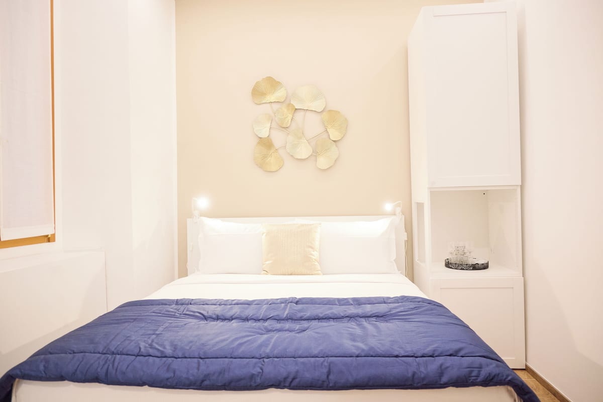 Ein ordentlich bezogenes Bett mit blauer Decke, weißen Kissen und einem dekorativen Wandbild darüber.