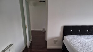 Un dormitorio con suelo de madera oscura, paredes blancas, cabecera negra y colcha blanca.