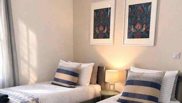 Une chambre double avec deux lits, chacun agrémenté d’une couvre-lit et de coussins à motifs, une table de chevet avec une lampe, et deux œuvres d’art encadrées accrochées au mur.