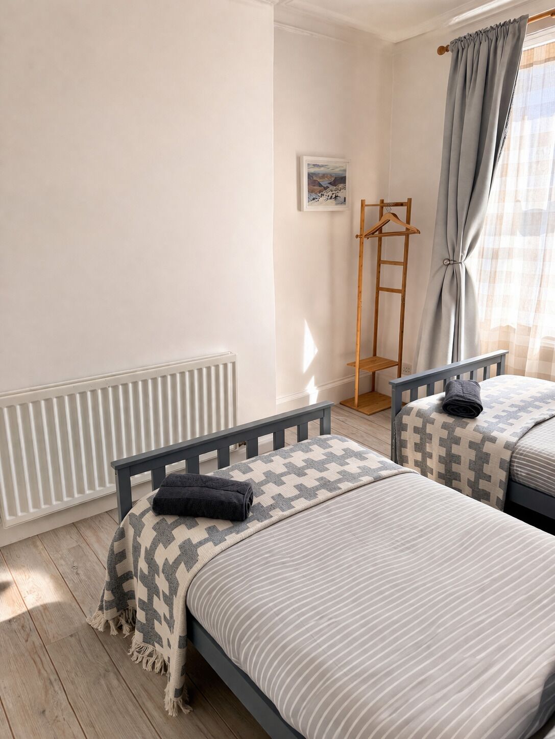A bedroom with two beds, a radiator, a wooden hanger, and a window with curtains.