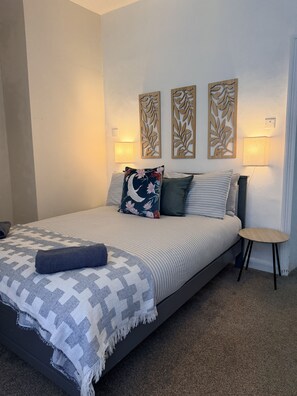 Una cama bien hecha con una colcha a rayas, un cojín decorativo y una manta. Hay dos lámparas fijadas a la pared y una mesita de noche con una lámpara al lado de la cama.