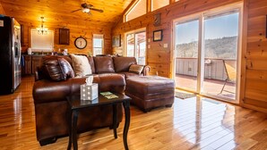 A cozy cabin interior with a wooden ceiling, a living area with a sofa and ottoman, a dining area with a clock, and large windows offering a view of the outdoors. - Charming riverside with stunning views, cozy interiors, full amenities, and spacious decks. (Draper)