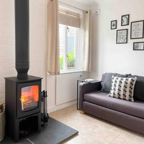 A living room with a black wood-burning stove, a grey sofa, and a window with curtains. - Cosy Annex sleeps 2 near Gretna, M6 Jct 45 / A75

 (Rigg)