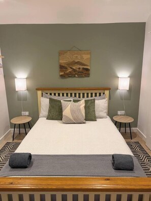 Una cama bien hecha con cabecera de madera, dos lámparas de noche y un cuadro sobre la cama.