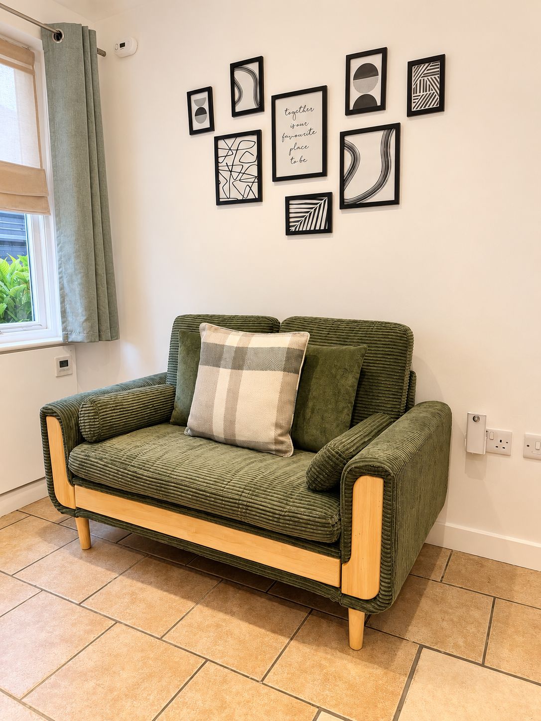 A green sofa with a plaid pillow, a window with curtains, and a wall with framed artwork.