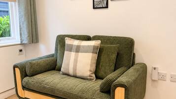 A green sofa with a plaid pillow, a window with curtains, and a wall with framed artwork.