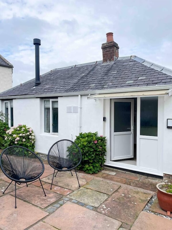 Une petite maison à l’extérieur blanc, avec un toit noir et une cheminée. On y voit deux chaises noires à l’extérieur et un pot de fleurs sur la droite.