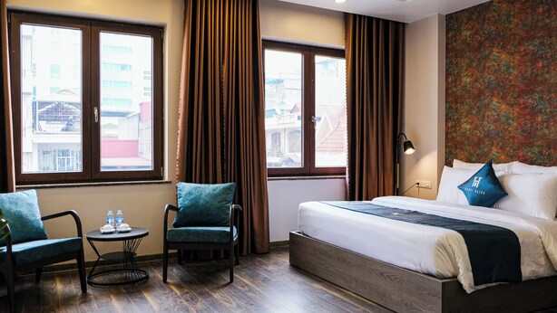 A hotel room with a large bed, two chairs, a small table with water bottles, and a view of buildings through large windows. - Hanoi Bliss Apartment Ba Dinh (Hanoi)