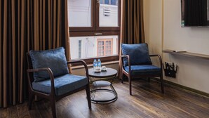 A hotel room with two blue armchairs, a small round table with water bottles, a TV mounted on the wall, and a view of a building through a window with curtains. - Hanoi Bliss Apartment Ba Dinh (Hanoi)
