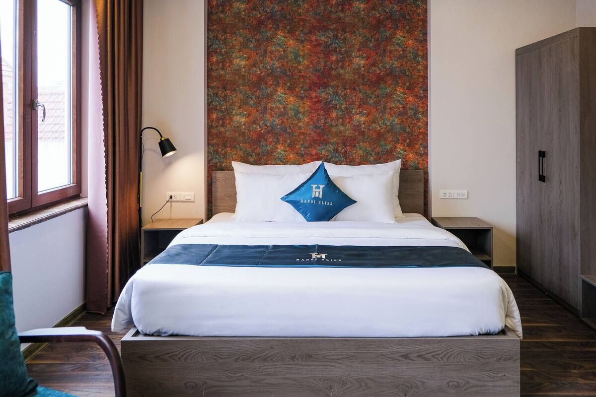 Ein Hotelzimmer mit einem großen Bett, einem Nachttisch, einer Lampe und Blick ins Freie.