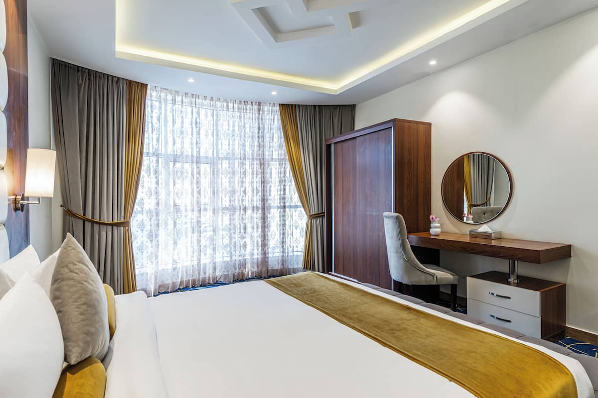 Habitación de hotel con una cama grande, un escritorio con silla, un armario y un ventanal con cortinas.